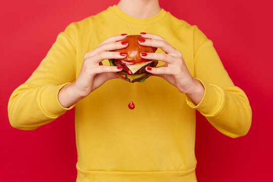 Woman eats burger as it drips tomato sauce. She wears a yellow jumper and stands against a red background.