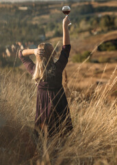 Young woman enjoying nature and sunlight in the field, the female is dressed in a purple dress and holding a glass with red wine