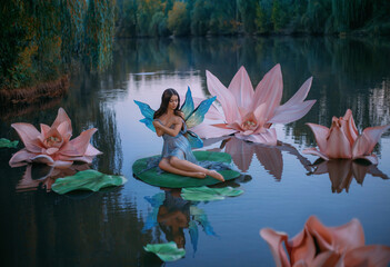 A beautiful woman a little fabulous fairy with butterfly wings sits on green water lily leaf. Fantasy scenery of huge pink flowers on the lake, green trees. River nymph innocent girl in an blue dress - obrazy, fototapety, plakaty