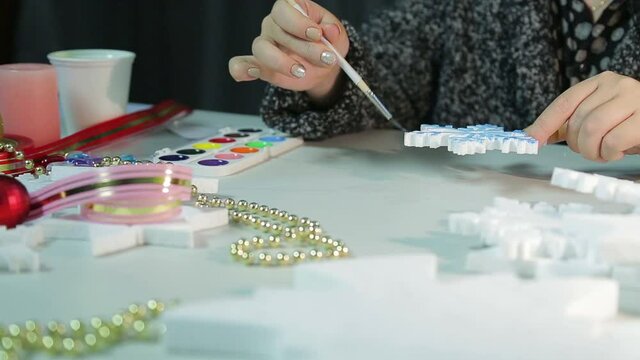 A young woman at the table makes home decorations for Christmas by painting white blanks with watercolors.