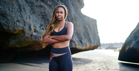 Fit ethnic young female standing on rocky coast in sportswear and looking at camera