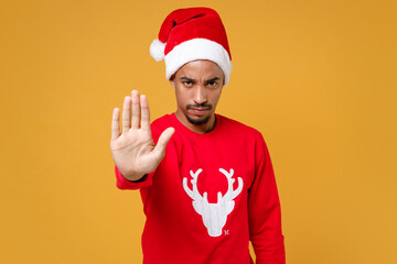 Fototapeta na wymiar Dissatisfied young Santa african american man 20s in red sweater Christmas hat showing stop gesture with palm isolated on yellow background studio portrait. Happy New Year celebration holiday concept.