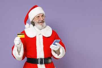 Fototapeta na wymiar Smiling Santa Claus man in Christmas hat red suit coat white gloves glasses using mobile phone hold credit bank card isolated on violet background. Happy New Year celebration merry holiday concept.