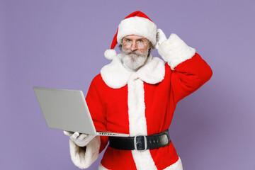 Puzzled Santa Claus man in Christmas hat red suit coat gloves glasses working on laptop pc computer put hand on head isolated on violet background. Happy New Year celebration merry holiday concept.