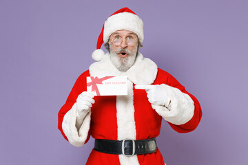 Fototapeta na wymiar Shocked elderly Santa Claus man in Christmas hat red suit coat gloves glasses pointing index finger on gift certificate isolated on violet background. Happy New Year celebration merry holiday concept.