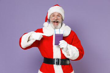 Fototapeta na wymiar Surprised traveler tourist Santa Claus man in Christmas hat red suit glasses pointing index finger on passport tickets isolated on violet background. Happy New Year celebration merry holiday concept.