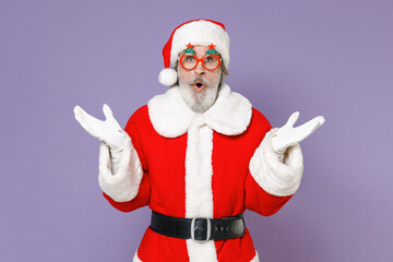 Fototapeta na wymiar Shocked Santa Claus man in Christmas hat red suit coat white gloves glasses keeping mouth open spreading hands isolated on violet background studio. Happy New Year celebration merry holiday concept.