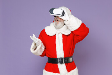 Fototapeta na wymiar Shocked Santa Claus man in Christmas hat red coat suit white gloves watching in vr headset gadget spreading hands isolated on violet background studio Happy New Year celebration merry holiday concept.