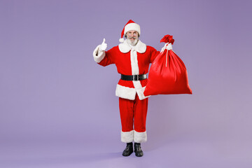 Full length portrait of funny Santa Claus man in Christmas hat coat suit hold bag with presents gifts showing thumb up isolated on violet background. Happy New Year celebration merry holiday concept.