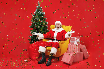 Surprised Santa Claus man in Christmas suit glasses sit in armchair with fir tree presents gifts confetti spreading hands isolated on red background. Happy New Year celebration merry holiday concept.