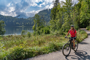 Fototapeta premium nice senior woman riding her electric mountain bike above the Freiberg Lake and a big ski flying hill in the Allgau Alps near Oberstdorf, Bavaria, Germany 