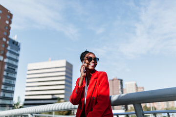 Enthusiastic black woman in red coat discussing on smartphone in downtown