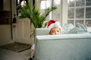 Cute young boy in a Santa hat hiding behind an arm rest of a big chair