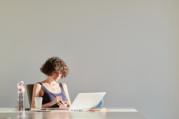 Casual curly woman writing in notepad sitting at desk in empty i