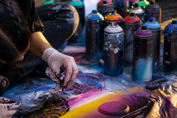 Fototapeta premium girl street artist in gloves paints with spray cans of paint on a piece of paper. multicolored cans of paint stand around. copy space