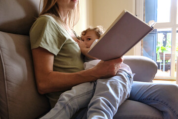 Mother breastfeeds her child while reading a relaxed book.