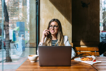 Happy businesswoman using laptop in cafeteria and talking on cellphone