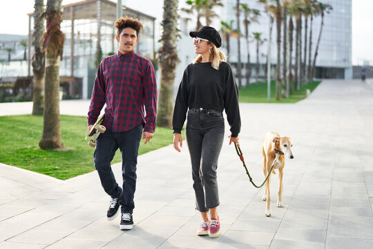 Stock photo of young couple in casual wear walking on city street with Greyhound dog on leash, man holding skateboard, trees and buildings on background. Owners and pet spending leisure outdoors