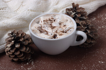 Cup of cocoa with marshmallows, white sweater, pine cones and snow on brown background. Autumn and winter concept. Chestnut color.