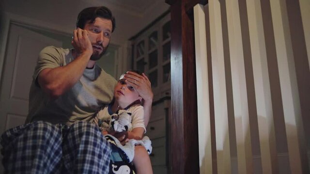 A father comforting his sick child at the top of a staircase while on the phone with a doctor, at bedtime