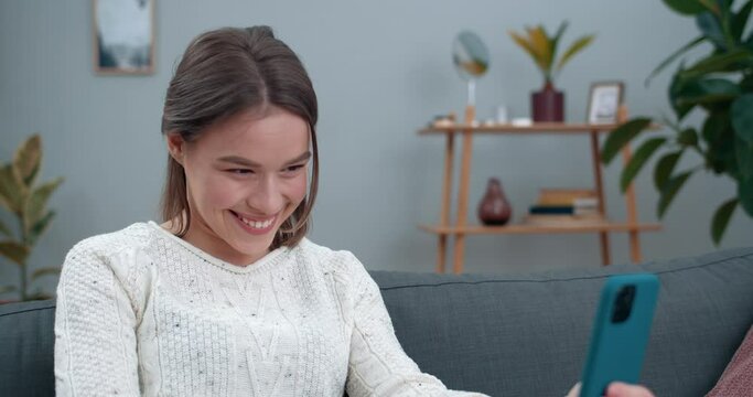 Crop view of woman having video call and showing with with deaf-mute sign language phrase I love you. Cheerful female person smiling and using smartphone while sitting on sofa.