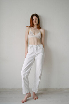 red-haired girl in white trousers and a white bra stands against a white wall