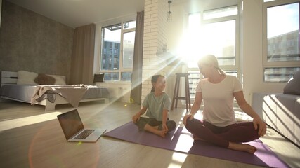 Mother and daughter practicing online yoga classes at home during the quarantine during the coronavirus pandemic. Family doing sports together online from home during isolation together at home
