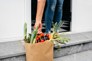 Anonymous woman picking up food delivery in paper bag from home porch