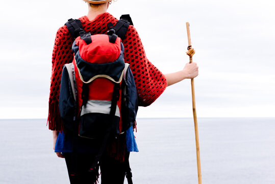 Cropped back view of unrecognizable female hiker with trekking stick standing against gray cloudy sky and enjoying freedom during pilgrimage through Spain