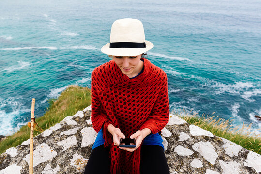 From above female backpacker with trekking stick sitting on stone border near sea and browsing smartphone while resting and checking route during pilgrimage through Spain