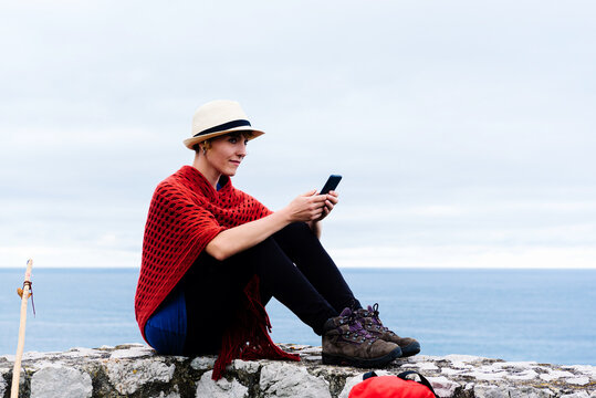 Full length side view of young female backpacker with trekking stick sitting on stone border near sea and browsing smartphone while resting and checking route during pilgrimage through Spain