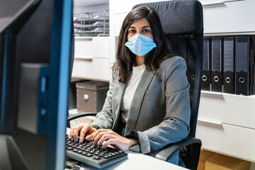 Confident young female specialist in formal suit and protective mask sitting at desk and using computer while working in modern workspace looking at camera