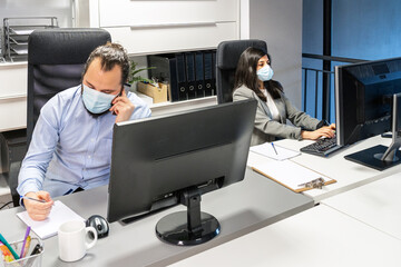 Serious focused young man in medical mask discussing business issue by phone while female colleague working with computer in contemporary coworking office