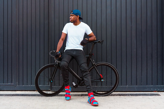 Full body of serious young African American man in casual street style wear and trendy boots standing with bike against gray wall on street and looking away