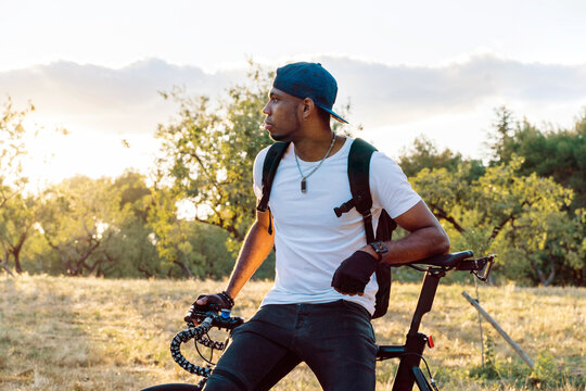 Calm young African American male in casual wear and cap with backpack leaning on bicycle and looking away while standing on green meadow against blurred hills in sunny summer day