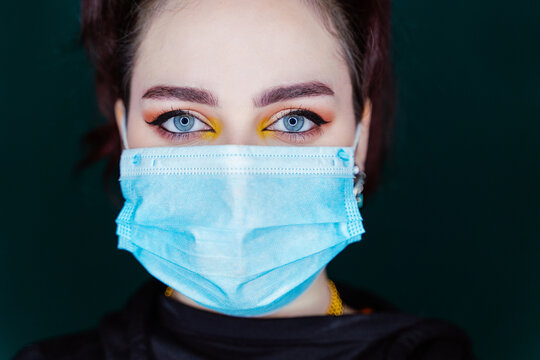 Calm female wearing medical mask standing in modern studio and looking at camera on dark background