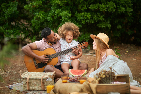 Happy young mixed race parents with cute curly haired daughter playing guitar and laughing while chilling together during picnic in summer day in green forest