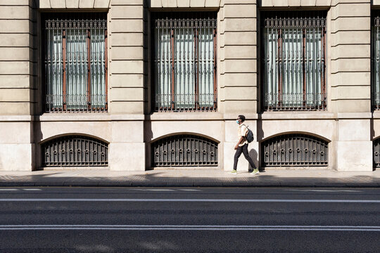 Side view of black male in medical mask walking along pavement near urban building on sunny day in Barcelona during coronavirus epidemic