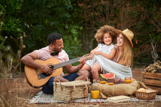 Happy young mixed race parents with cute curly haired daughter playing guitar and laughing while chilling together during picnic in summer day in green forest