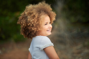 Adorable little girl with blonde Afro hair smiling and looking away while spending summer day in nature