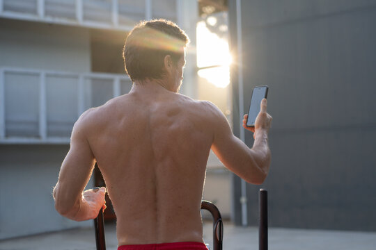 Back view of athletic male with naked torso doing exercises on stepper and taking selfie on smartphone during workout in yard