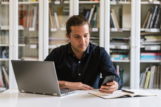 Focused businessman in formal shirt sitting at table while doing paperwork and typing on notebook in bright workspace