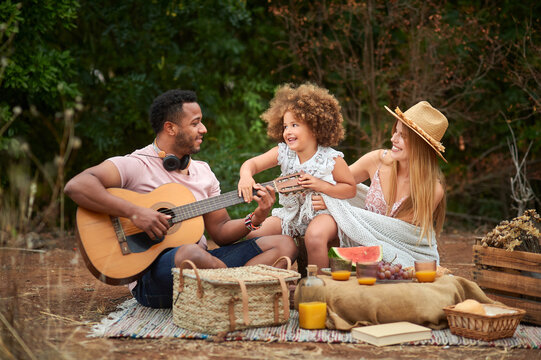 Happy young mixed race parents with cute curly haired daughter playing guitar and laughing while chilling together during picnic in summer day in green forest
