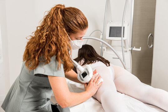 Masseuse in protective mask doing anti cellulite lipo massage to female client dressed in white suit during body care procedure in salon of apparatus cosmetology