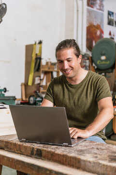 Content male carpenter sitting at workbench and working on netbook on project in modern workshop