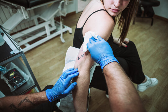 Back view crop anonymous male tattooist in latex gloves wiping freshly made tattoo on arm of female client with napkin while working in salon