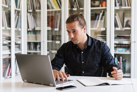 Focused businessman in formal shirt sitting at table while doing paperwork and typing on notebook in bright workspace
