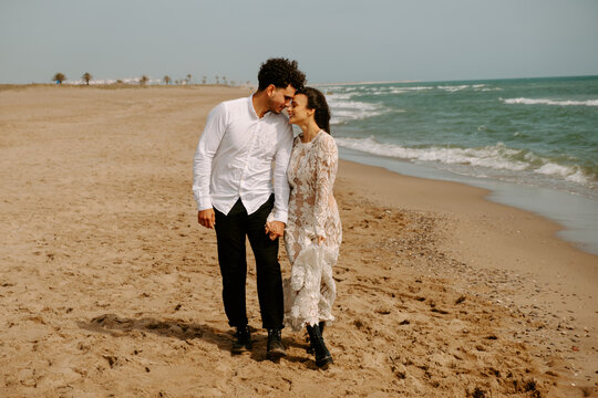 Full length happy young bride and groom in elegant boho outfits holding hands and embracing each other with eyes closed while strolling along sandy beach with waving sea