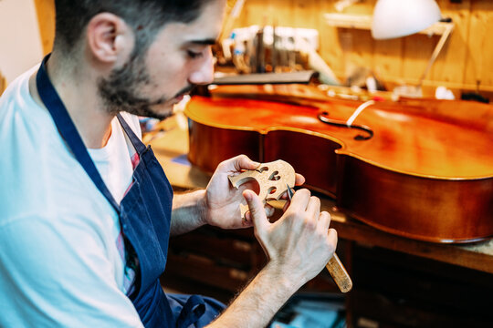 Side view of skilled young male artisan with knife carving wooden violin bridge while creating string instrument in workshop