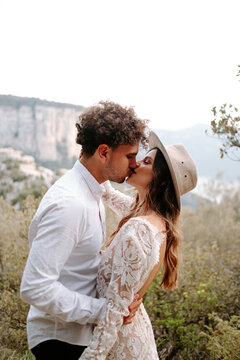 Side view of romantic young newly married couple in wedding outfits with bouquet embracing and kissing while standing against picturesque mountain landscape of Morro de Labella in Spain